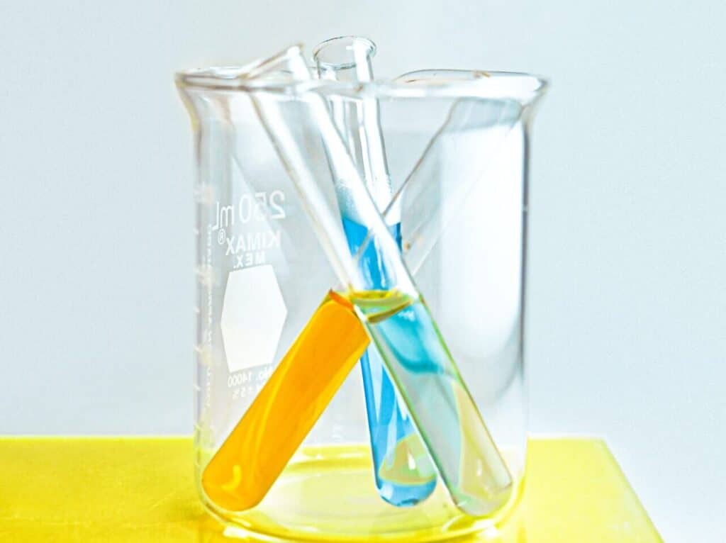 Three glass test tubes, with yellow, blue and clear liquid, sitting inside of a glass beaker.